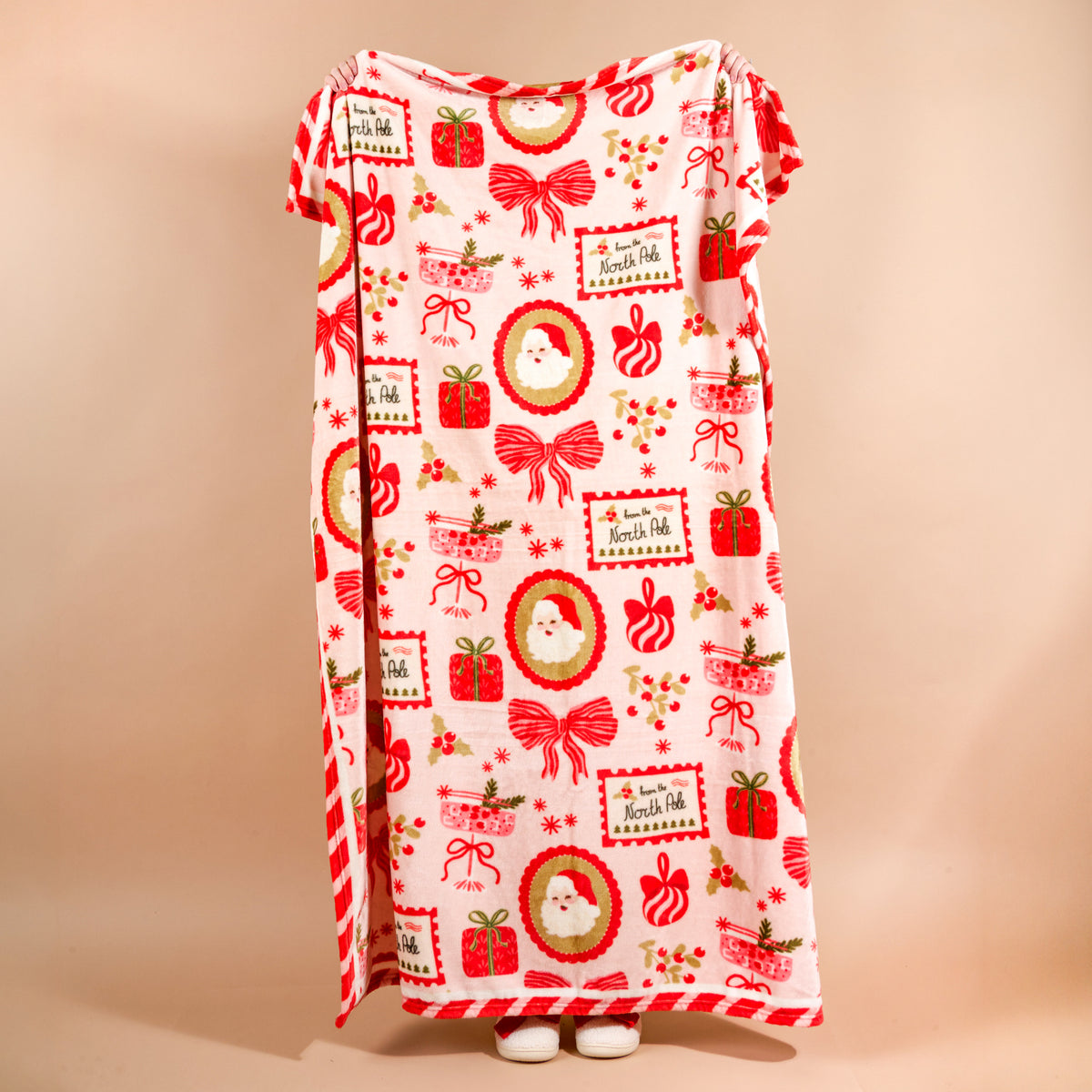 A person holds up a Mistletoe Magic Snuggle Up Plush Blanket featuring a cozy pattern of Santa Claus faces, red gift boxes, bows, and From the North Pole labels.