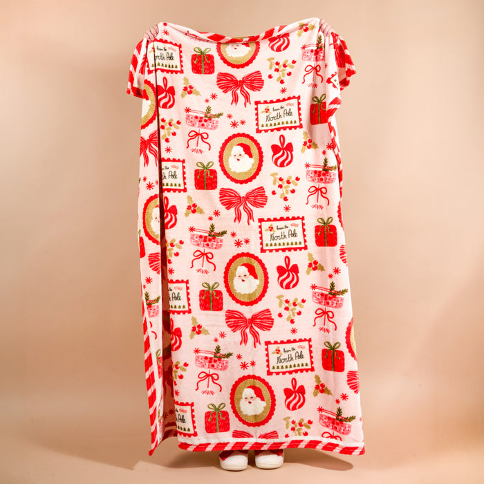 A person holds up a Mistletoe Magic Snuggle Up Plush Blanket featuring a cozy pattern of Santa Claus faces, red gift boxes, bows, and From the North Pole labels.
