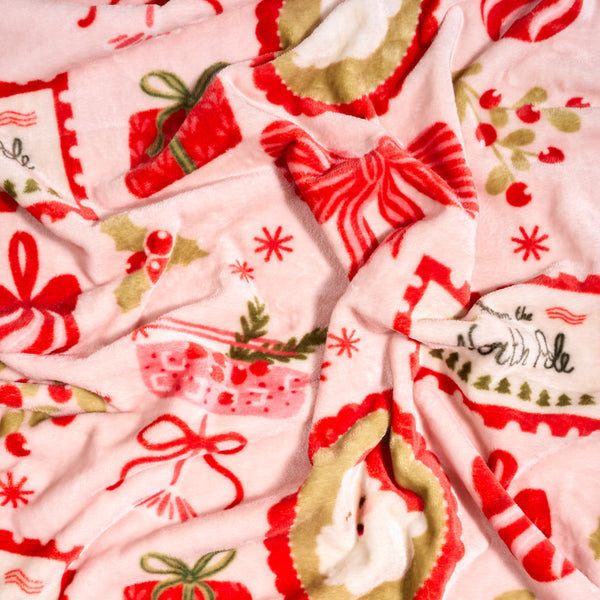 Mistletoe Magic Snuggle Up Plush Blanket in soft, light pink featuring a pattern of Christmas gifts, bows, holly, snowflakes, and a North Pole stamp design.