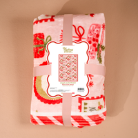 The Darling Effect Mistletoe Magic Snuggle Up Plush Blanket, folded and featuring a pink and white Christmas-themed pattern of presents and wreaths, secured with a branded paper sleeve.