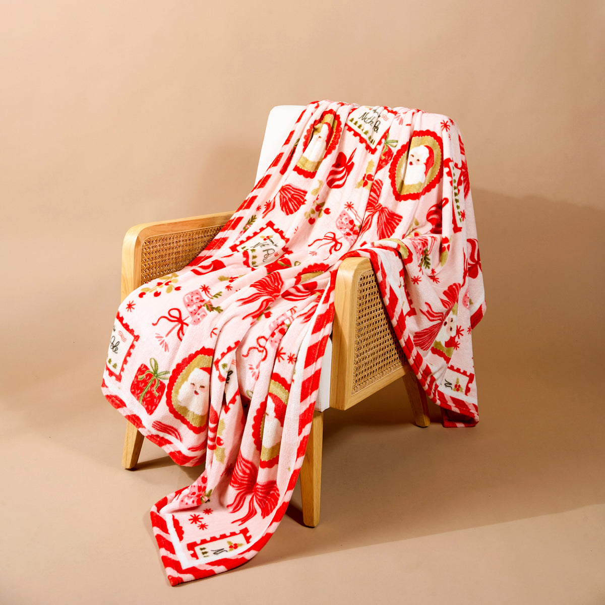 Mistletoe Magic Snuggle Up Plush Blanket in light pink with a red Christmas print of Santa, presents, and candy cane stripes, draped over a wooden chair.