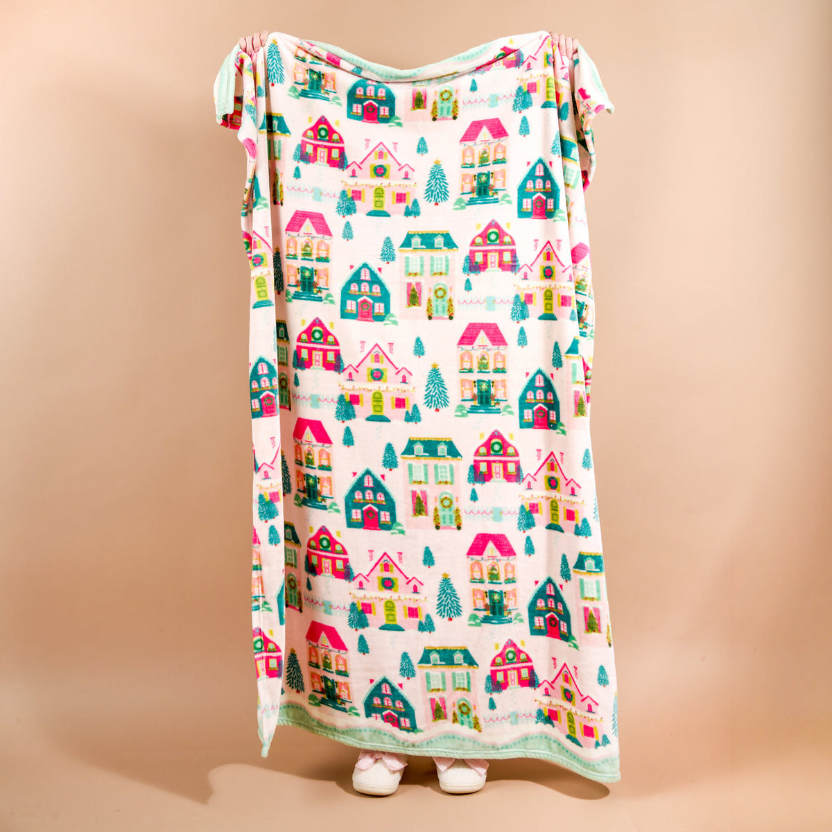 A person holds up a cozy Sugarplum Lane Snuggle Up Plush Blanket by The Darling Effect, decorated with a pattern of colorful houses and pine trees.