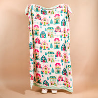 A person holds up a cozy Sugarplum Lane Snuggle Up Plush Blanket by The Darling Effect, decorated with a pattern of colorful houses and pine trees.