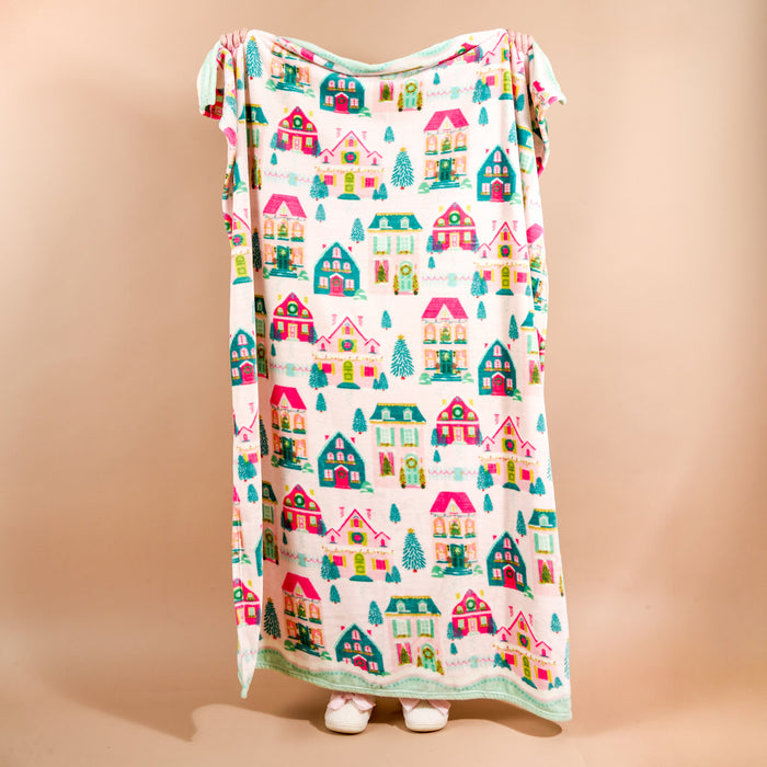 A person holds up a cozy Sugarplum Lane Snuggle Up Plush Blanket by The Darling Effect, decorated with a pattern of colorful houses and pine trees.