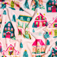 Sugarplum Lane Snuggle Up Plush Blanket featuring a repeating pattern of colorful holiday houses, wreaths, and pine trees on a light pink background.