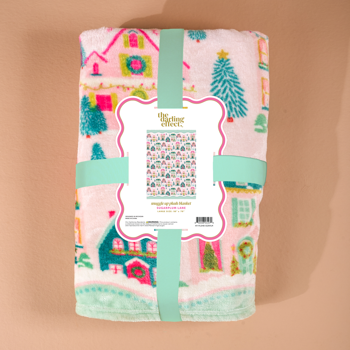 Sugarplum Lane Snuggle Up Plush Blanket by The Darling Effect, shown as a folded, velvety blanket with a repeating pattern of colorful holiday houses and pine trees, secured with a branded paper band.