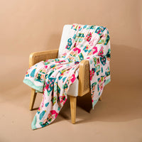 A Sugarplum Lane Snuggle Up Plush Blanket with colorful houses and trees draped over a wooden armchair.