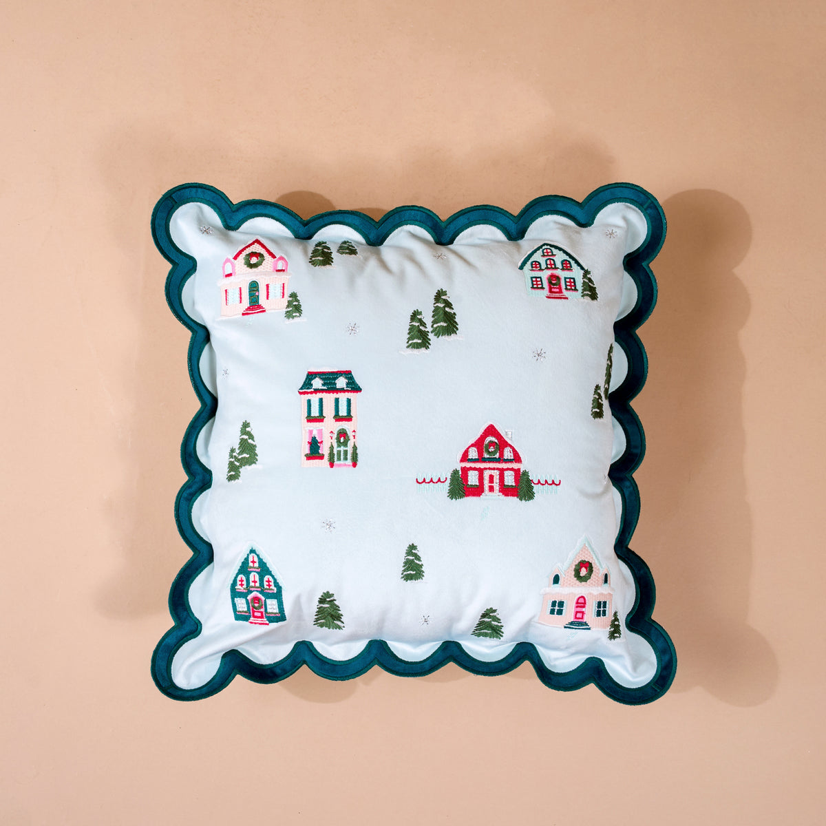 Home Sweet Home Scalloped Square Throw Pillow: a square light blue decorative pillow with a scalloped dark green border, featuring an embroidered pattern of winter houses, Christmas wreaths, and evergreen trees.