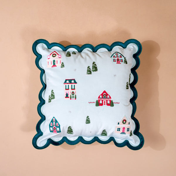 Home Sweet Home Scalloped Square Throw Pillow: a square light blue decorative pillow with a scalloped dark green border, featuring an embroidered pattern of winter houses, Christmas wreaths, and evergreen trees.