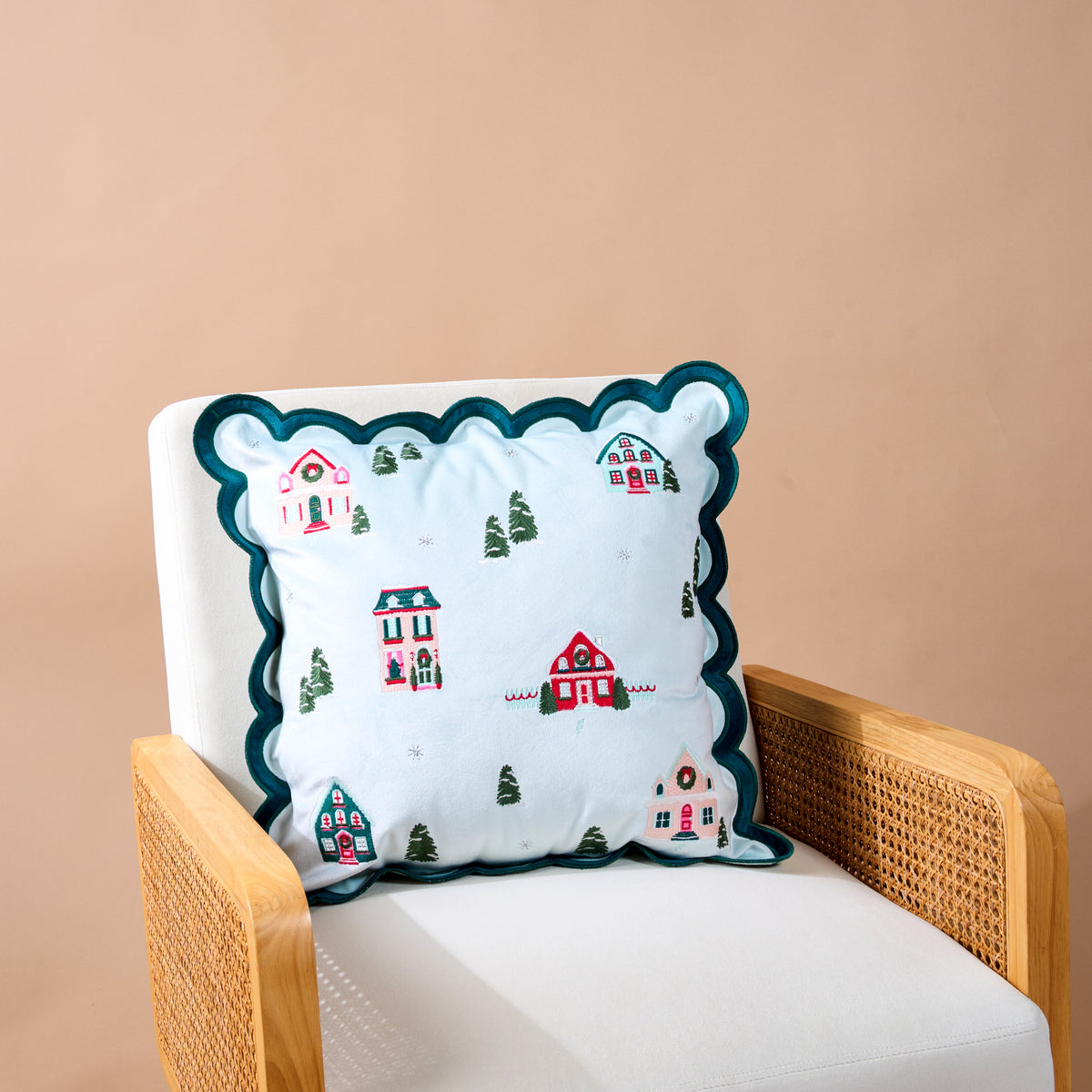 Home Sweet Home Scalloped Square Throw Pillow with a dark green scalloped edge and zippered closure, featuring an embroidered winter village scene on a light blue background, resting on a white armchair.
