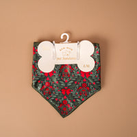 A Merry Moment Bow-Wow Pet Bandana made of satin featuring a dark green pattern of red berries, bows, and holly leaves, attached to a bone-shaped cardboard display card.
