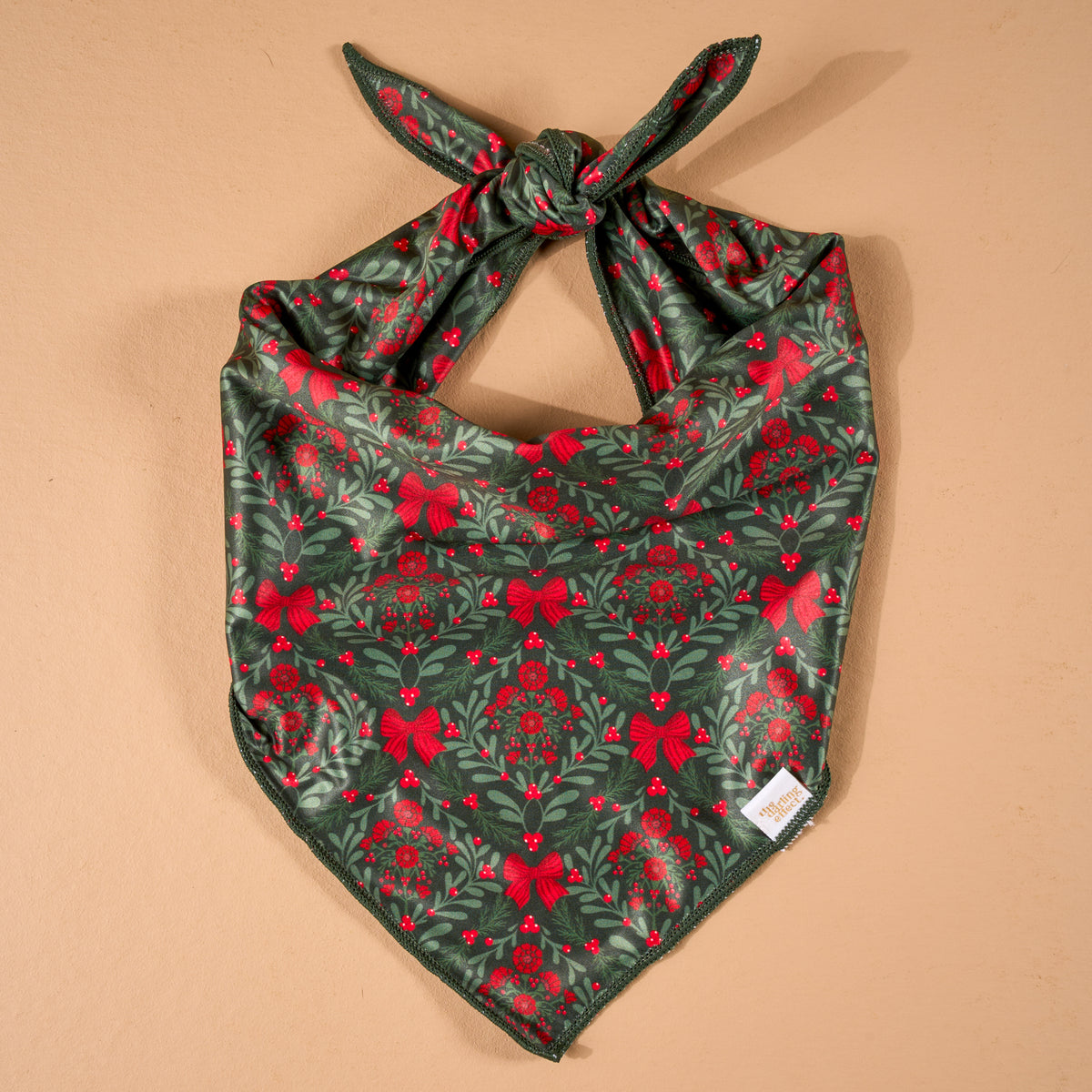 A Merry Moment Bow-Wow Pet Bandana, a triangular satin pet bandana with a dark green pattern of red bows, mistletoe, and berries, shown tied in a knot at the top.