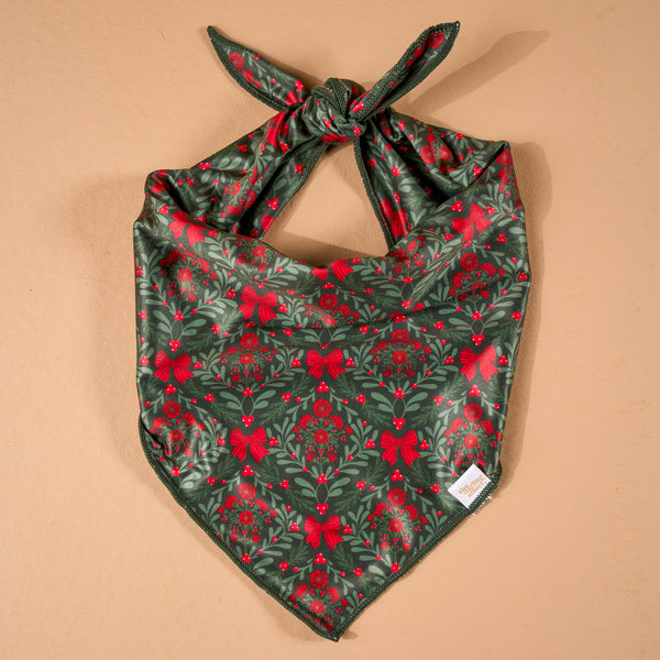 A Merry Moment Bow-Wow Pet Bandana, a triangular satin pet bandana with a dark green pattern of red bows, mistletoe, and berries, shown tied in a knot at the top.