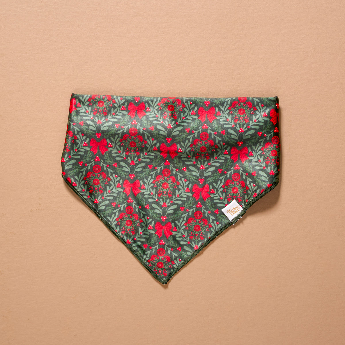 A Merry Moment Bow-Wow Pet Bandana featuring a triangular festive pattern of green mistletoe, red berries, and red bows on a dark green background.