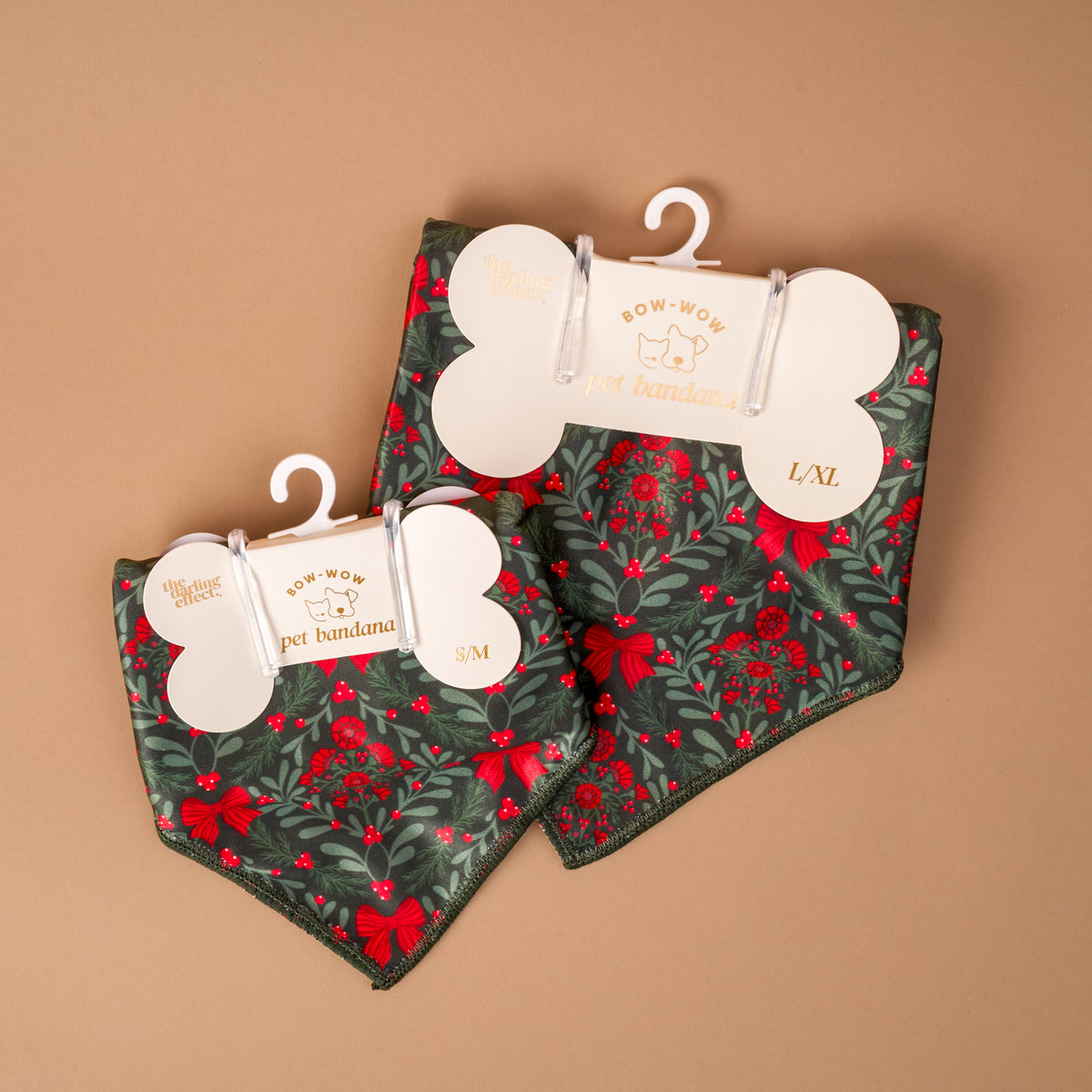 A Merry Moment Bow-Wow Pet Bandana set featuring two triangular bandanas with a dark green pattern of red ribbons, holly berries, and evergreen sprigs, packaged on bone-shaped cardboard hangers.