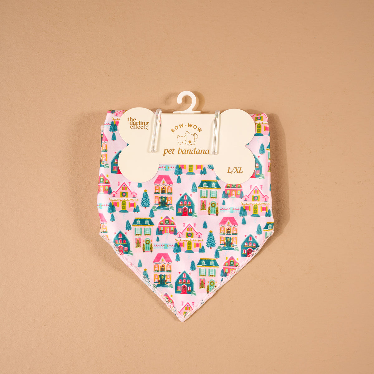 Sugarplum Lane Bow-Wow Pet Bandana in pink satin, featuring a repeating pattern of colorful holiday houses and evergreen trees, hanging from a bone-shaped retail card.