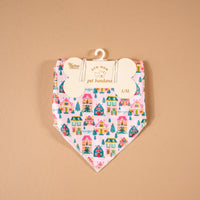Sugarplum Lane Bow-Wow Pet Bandana in pink satin, featuring a repeating pattern of colorful holiday houses and evergreen trees, hanging from a bone-shaped retail card.