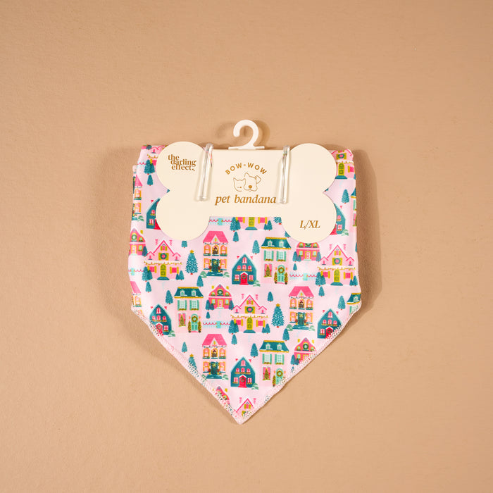 Sugarplum Lane Bow-Wow Pet Bandana in pink satin, featuring a repeating pattern of colorful holiday houses and evergreen trees, hanging from a bone-shaped retail card.
