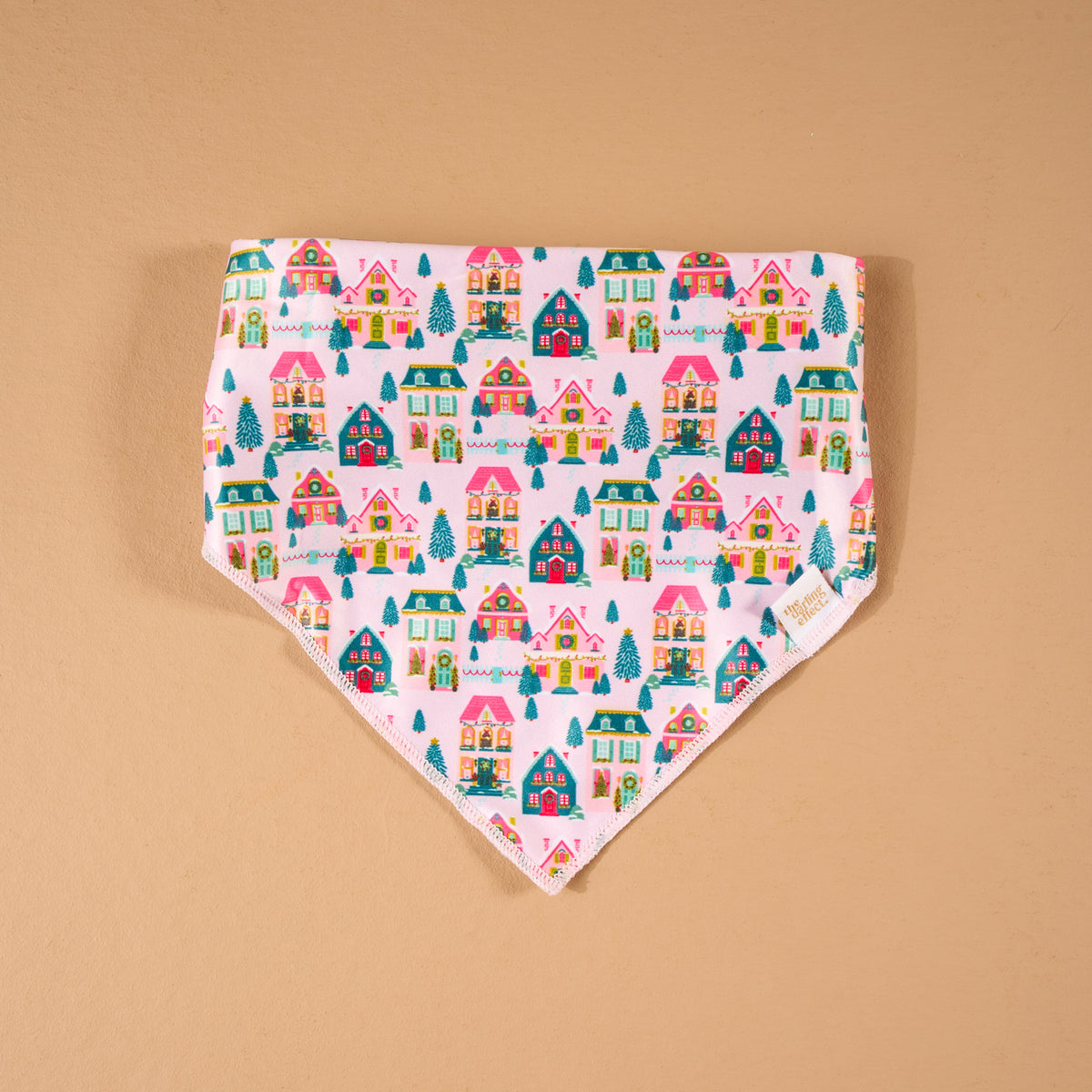 Sugarplum Lane Bow-Wow Pet Bandana, a triangular satin pet bandana featuring a pattern of colorful illustrated houses and pine trees on a light pink background.