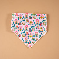 Sugarplum Lane Bow-Wow Pet Bandana, a triangular satin pet bandana featuring a pattern of colorful illustrated houses and pine trees on a light pink background.