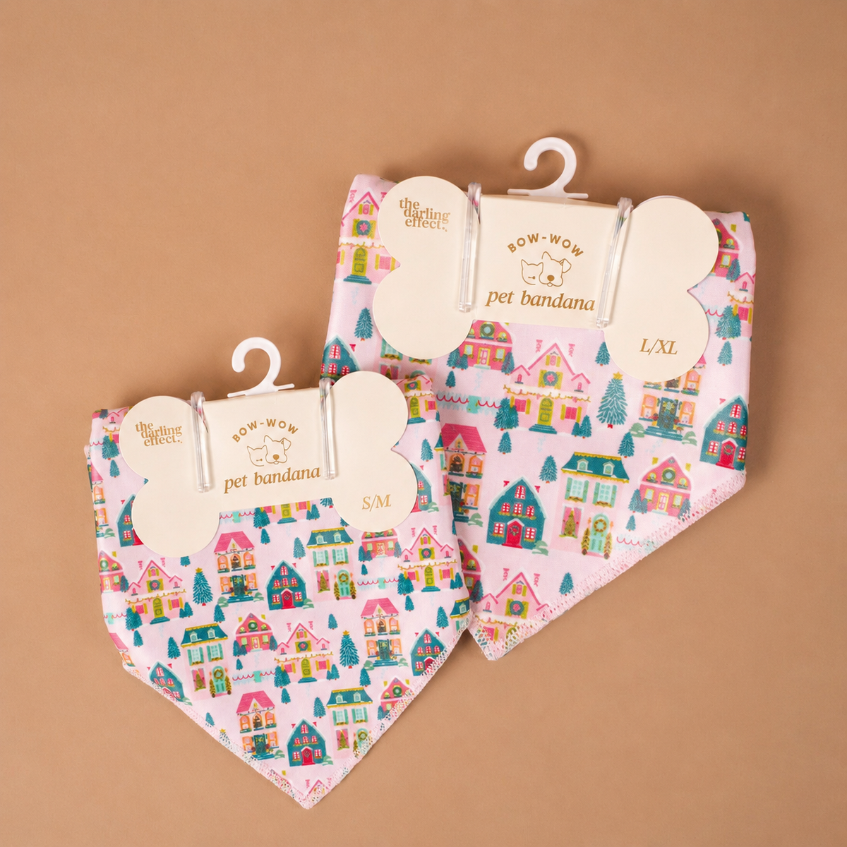 Two triangular Sugarplum Lane Bow-Wow Pet Bandanas in satin with a pink holiday village print, each displayed on a bone-shaped hanger.