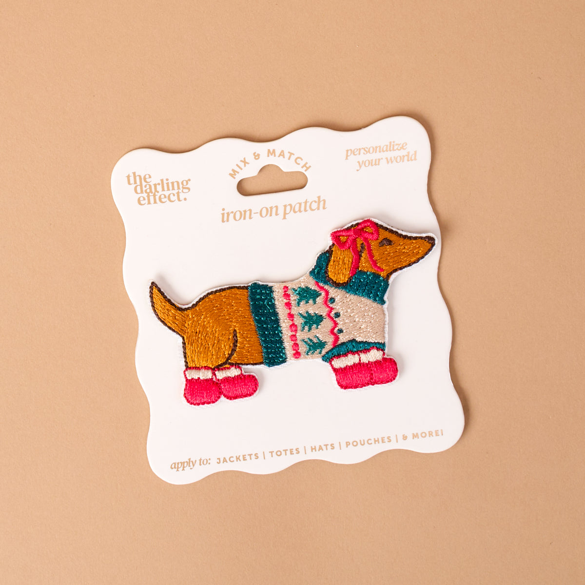 Dashing Dachshund Mix & Match Iron-On Patch by The Darling Effect featuring an embroidered dachshund with colorful designs, a festive sweater, a pink head bow, and pink booties, displayed on a backing card.