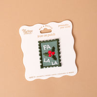 Fa La La Mix & Match Iron-On Patch displayed on a product card, featuring a rectangular, stamp-shaped design with the text FA LA LA and a sprig of holly with red berries on a green and white background.