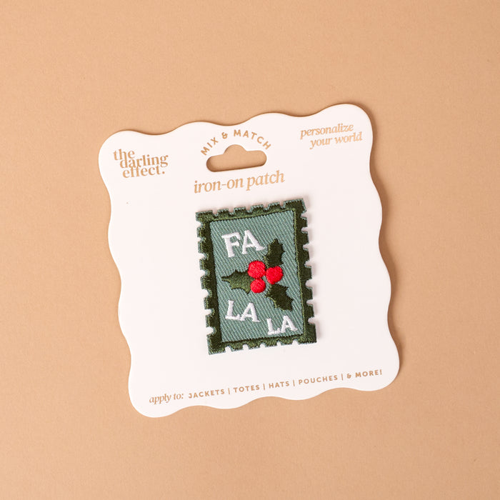 Fa La La Mix & Match Iron-On Patch displayed on a product card, featuring a rectangular, stamp-shaped design with the text FA LA LA and a sprig of holly with red berries on a green and white background.