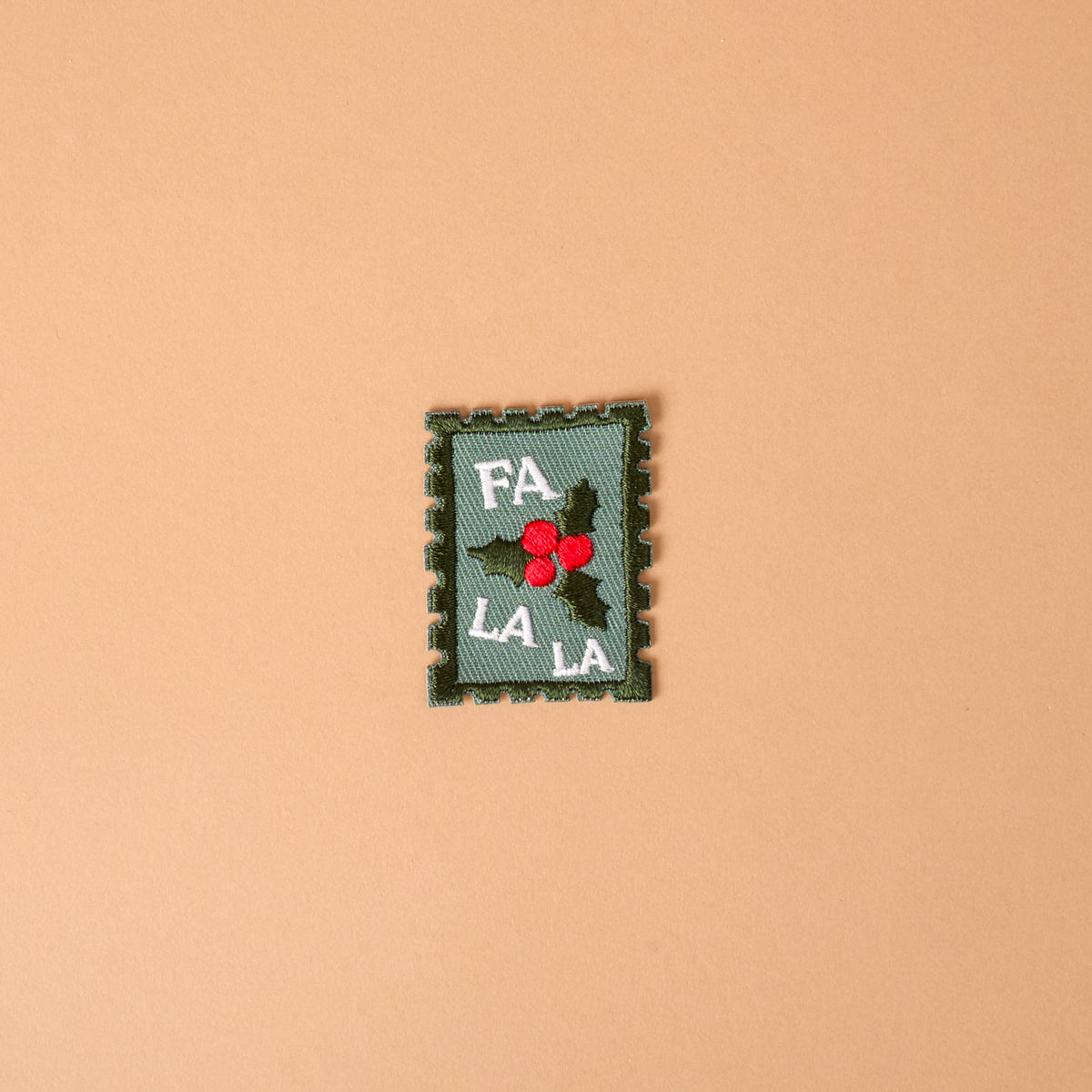 Fa La La Mix & Match Iron-On Patch featuring a rectangular, postage stamp-shaped design with green holly leaves, red berries, and the text FA LA LA.
