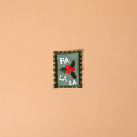 Fa La La Mix & Match Iron-On Patch featuring a rectangular, postage stamp-shaped design with green holly leaves, red berries, and the text FA LA LA.