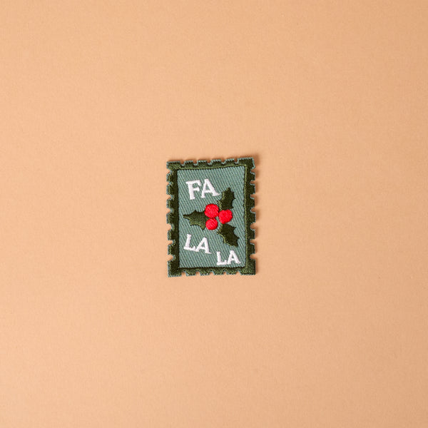 Fa La La Mix & Match Iron-On Patch featuring a rectangular, postage stamp-shaped design with green holly leaves, red berries, and the text FA LA LA.