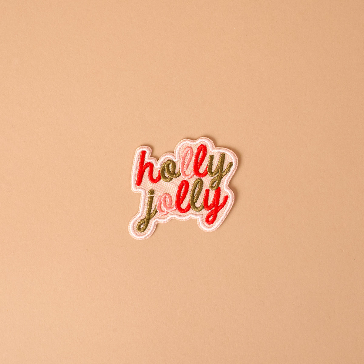 The Holly Jolly Mix & Match Iron-On Patch is a speech bubble shaped patch featuring the words holly jolly embroidered in red and gold script.