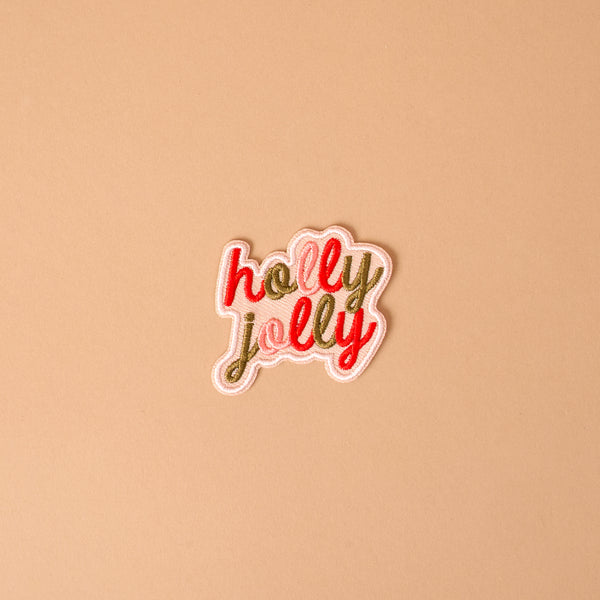 The Holly Jolly Mix & Match Iron-On Patch is a speech bubble shaped patch featuring the words holly jolly embroidered in red and gold script.