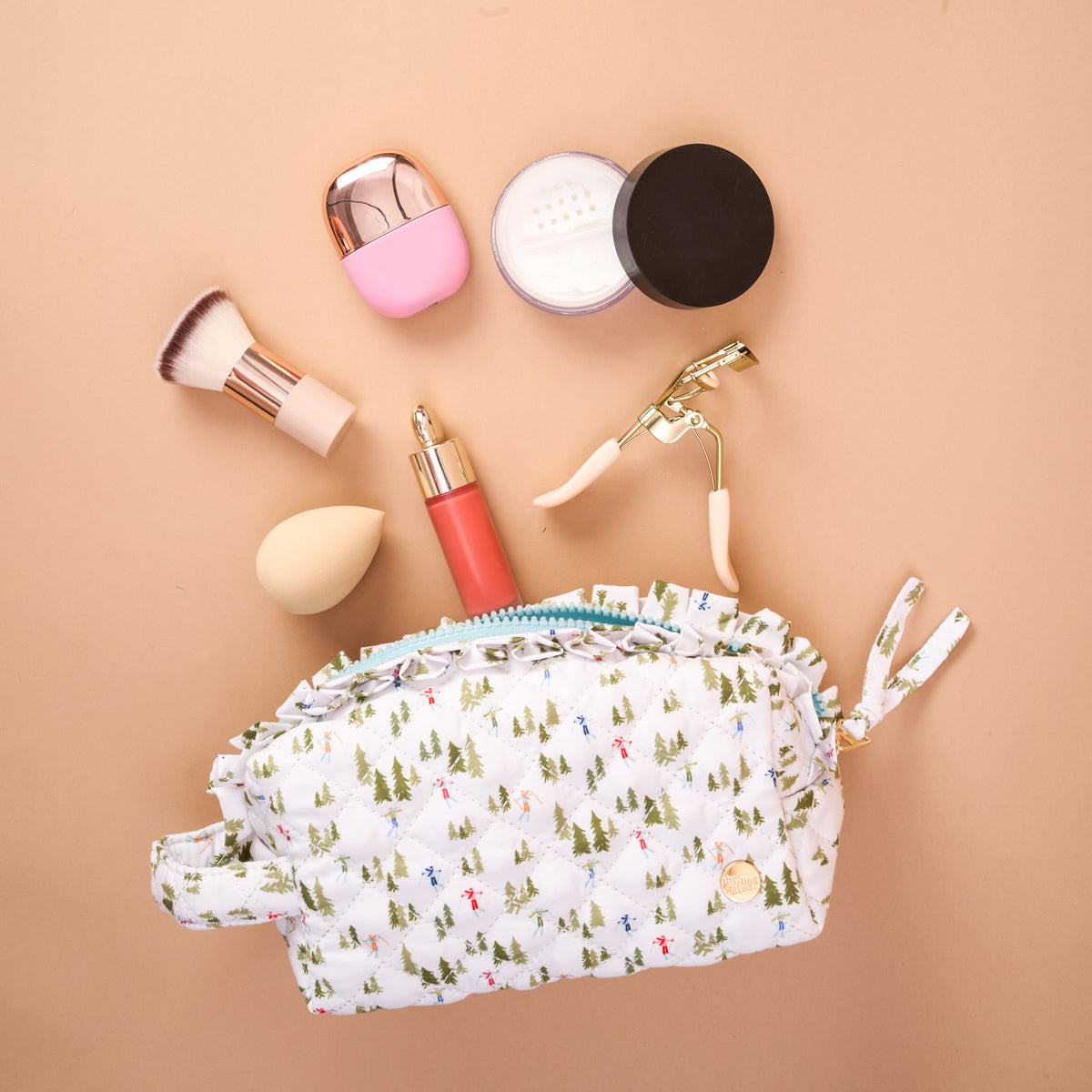 An Aprés Ski Essentials Quilted Cosmetic Pouch patterned with skiers and trees, surrounded by makeup brushes, a sponge, an eyelash curler, and various beauty products.