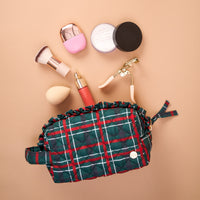 An open green and red plaid Evergreen Plaid Essentials Quilted Cosmetic Pouch with makeup brushes, a beauty sponge, a powder container, a serum bottle, and an eyelash curler arranged above it.