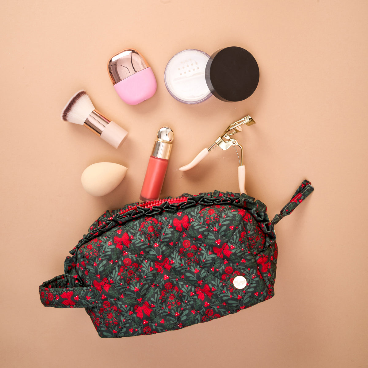 A Merry Moment Essentials Quilted Cosmetic Pouch, an open, quilted dark green cosmetic pouch with a red floral pattern, is displayed with makeup brushes, a sponge, a lipstick, an eyelash curler, and loose powder arranged above it.