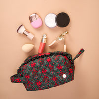 A Merry Moment Essentials Quilted Cosmetic Pouch, an open, quilted dark green cosmetic pouch with a red floral pattern, is displayed with makeup brushes, a sponge, a lipstick, an eyelash curler, and loose powder arranged above it.