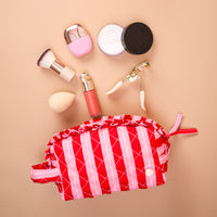 The Darling Effect Peppermint Stripes Essentials Quilted Cosmetic Pouch, featuring red and pink stripes and a ruffle trim, shown with a makeup sponge, brush, eyelash curler, and various cosmetic containers inside.