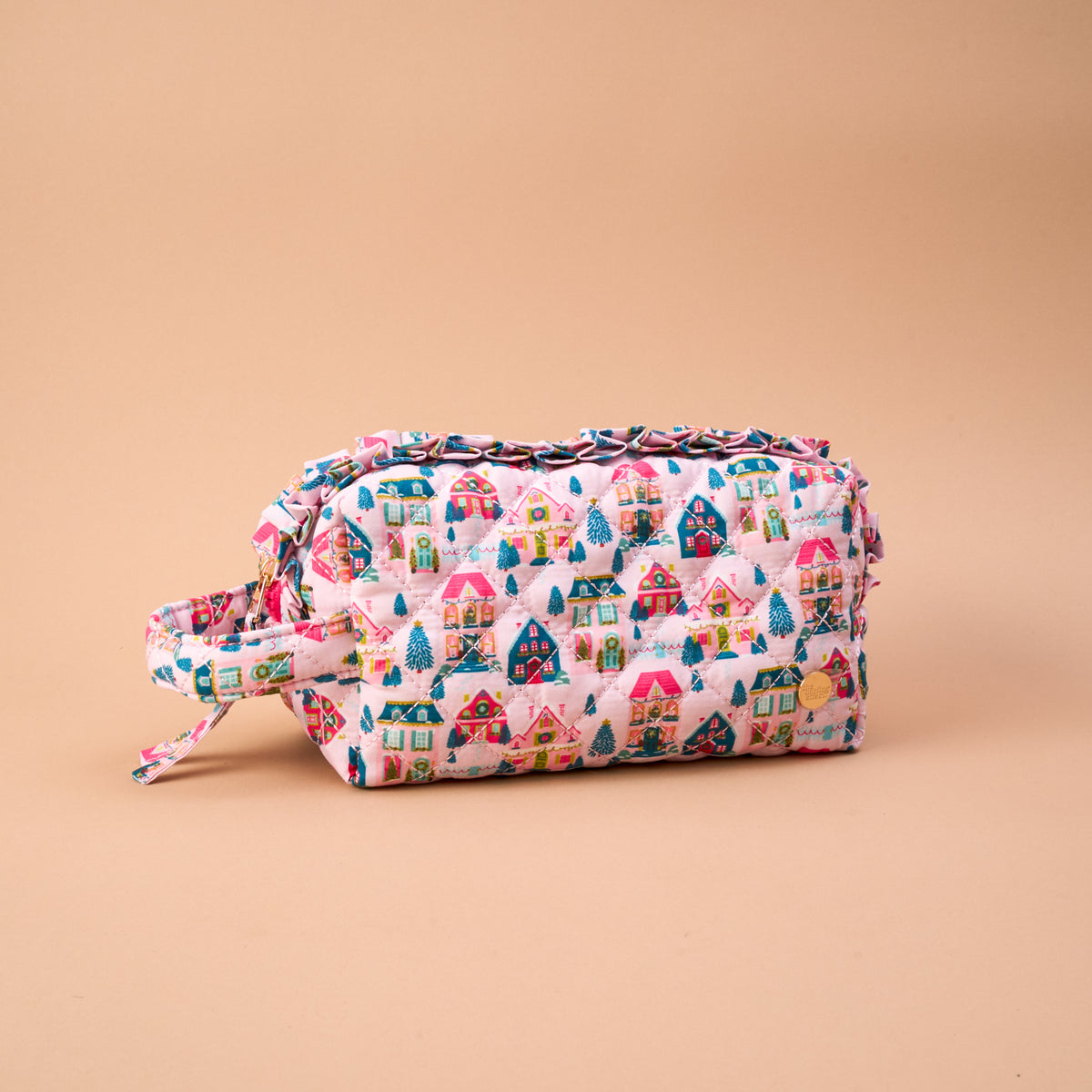 Sugarplum Lane Essentials Quilted Cosmetic Pouch in pink with a print of colorful holiday houses and pine trees, featuring ruffled trim and a side carry handle.