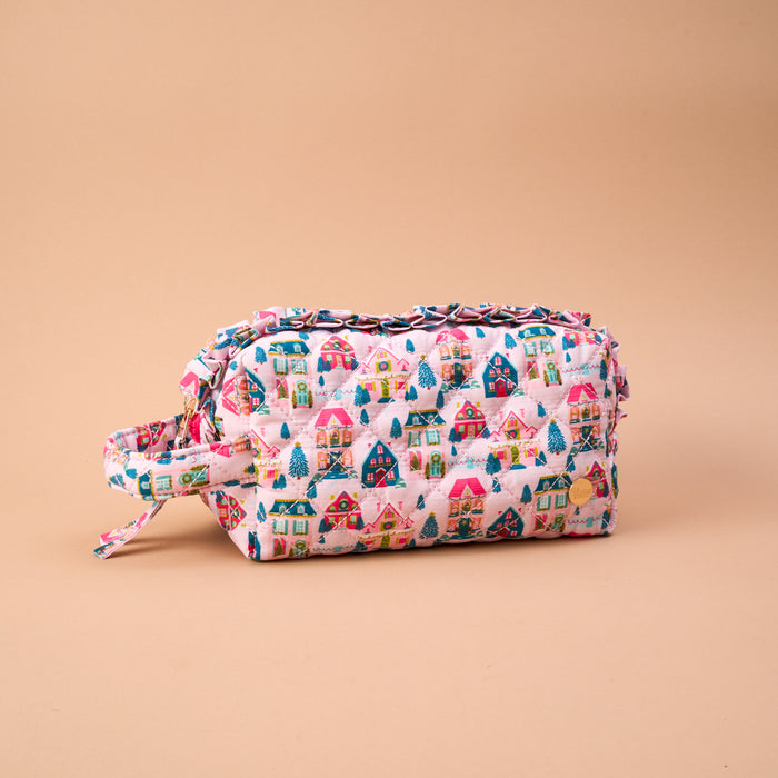 Sugarplum Lane Essentials Quilted Cosmetic Pouch in pink with a print of colorful holiday houses and pine trees, featuring ruffled trim and a side carry handle.