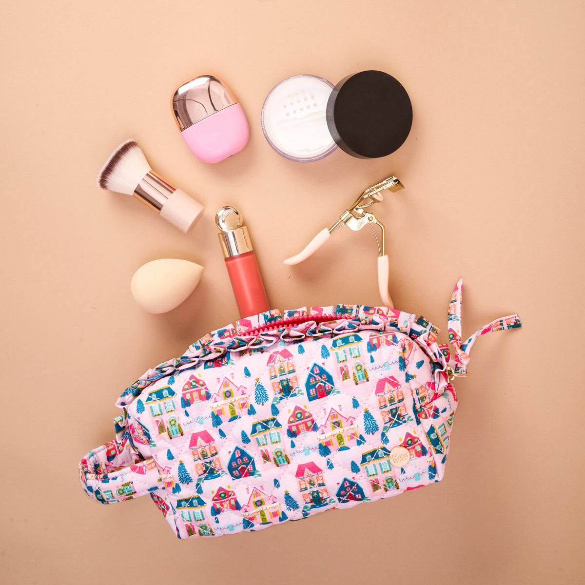 A Sugarplum Lane Essentials Quilted Cosmetic Pouch with a colorful house pattern, surrounded by a makeup brush, sponge, eyelash curler, powder container, and lip gloss.