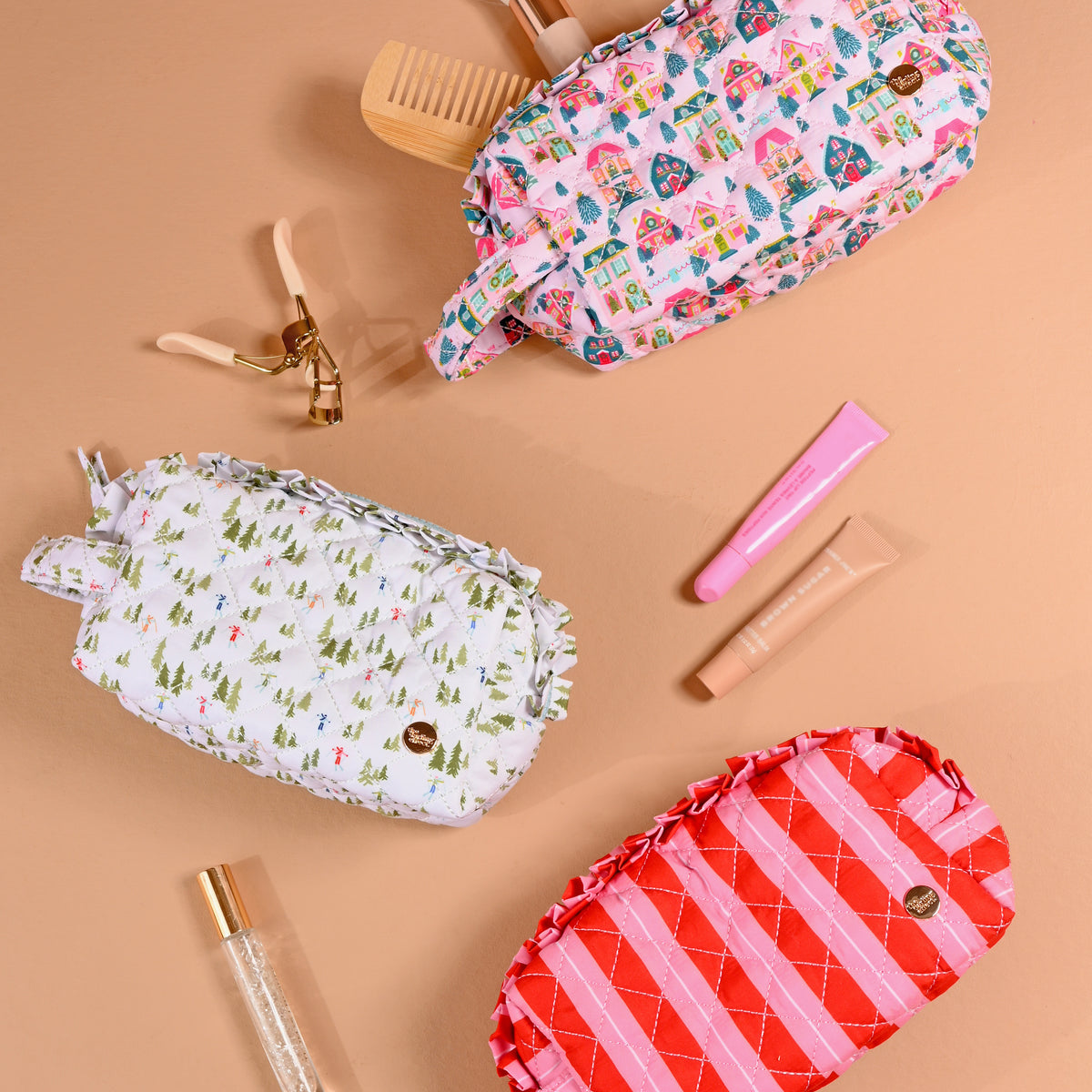 Three Sugarplum Lane Essentials Quilted Cosmetic Pouch bags with ruffled edges featuring prints of holiday houses, winter trees, and red and pink stripes, arranged with various beauty accessories on a tan surface.