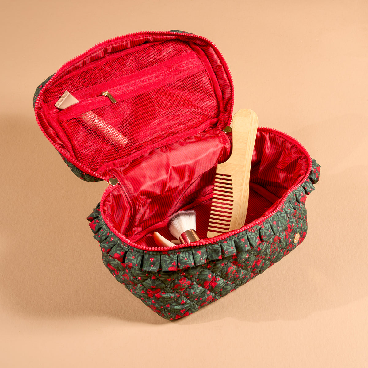 Open quilted dark green A Merry Moment Glam & Go Quilted Cosmetic Bag with a red holly print and a ruffled trim, containing a wooden comb and makeup brushes.