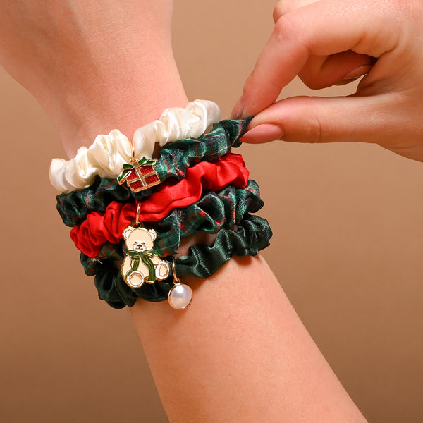A hand wears the Evergreen Plaid Scrunchie Charm Set, featuring four scrunchies in cream, red, and green plaid, accented with gift box, teddy bear, and pearl bead charms.