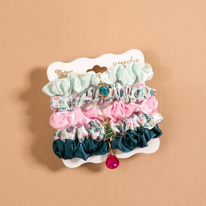 The Sugarplum Lane Scrunchie Charm Set includes four scrunchies on a display card, featuring light green, pink, dark green, and two patterned designs with teal teardrop, Christmas tree, and pink teardrop charms.