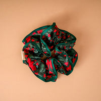 A Merry Moment Ruffle Up Scrunchie in dark green with a festive pattern of red berries and pine needles, featuring a solid green ruffled edge.