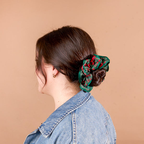 A Merry Moment Ruffle Up Scrunchie: an oversized, green, snag-free hair accessory featuring a red berry and leaf pattern, worn in a hair bun.