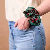 A Merry Moment Ruffle Up Scrunchie by The Darling Effect, featuring an oversized, ruffled design with a festive pattern of red berries and green leaves worn around a wrist.