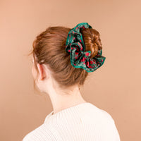 A person wears an oversized, dark green A Merry Moment Ruffle Up Scrunchie by The Darling Effect, featuring a red floral pattern, in a bun.
