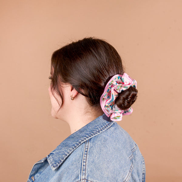 A person wearing a hair bun held by a Sugarplum Lane Ruffle Up Scrunchie featuring an oversized, colorful patterned ruffle design.